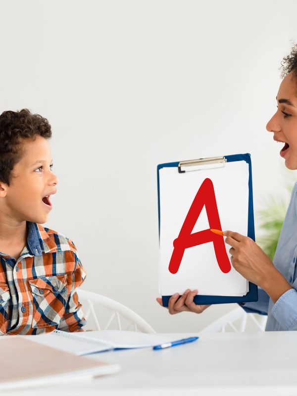 Cheerful speech therapist curing boy's problems and impediments, showing paper with A letter, kid learning and pronouncing letters with private English language tutor during lesson, sitting indoor