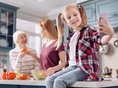 Domestic moment. Adorable little girl sitting on the kitchen counter and taking selfies while her mother and grandmother cooking salad in the background